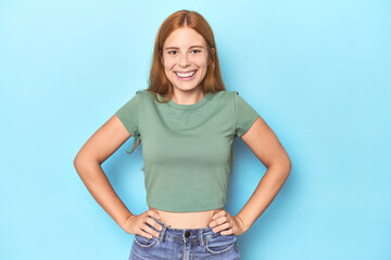 Wall Mural - Redhead young woman on blue background confident keeping hands on hips.