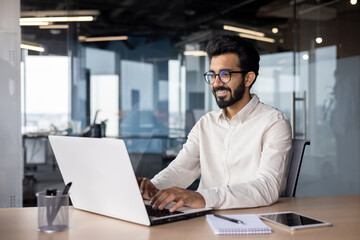 Wall Mural - A young smiling Indian man is working in the office on a laptop, texting, chatting