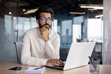 Portrait of a thoughtful and serious Indian male businessman sitting in the office at the workplace and working on a laptop