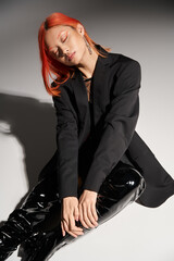 fashionable asian woman in sexy latex boots and blazer sitting with closed eyes on grey backdrop