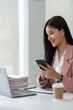 © crizzystudio - Young Asian businesswoman using mobile phone surfing the internet searching for financial information. Statistics, graphs, income charts. while working online on laptop computer at office..
