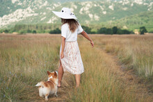 Happy Girl Walk With Dog. Pembroke Corgi With Woman In Hat On Nature On The Field In Summer