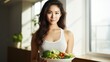 Asian woman is holding a salad bowl and looking at the camera. A Beautiful girl in sportswear likes to eat clean vegetables after exercising for a healthy home. Diet and healthy food concept
