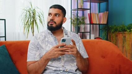 Indian man sits on couch uses mobile phone smiles at home living room apartment. Young Arabian guy texting share messages content on smartphone social media applications online, watching relax movie