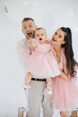 Wall Mural - Cheerful spouse in stylish clothes hugging with love pretty baby girl at decorated studio. Joyful family of father, mother and little daughter sharing happy moments of birthday celebration.