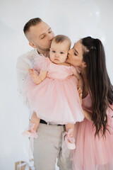 Wall Mural - Cheerful young parents kissing little daughter wearing festive pink dress and creating precious memories during first anniversary in white studio. Concept of love, holiday and celebration.