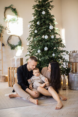 Wall Mural - Delighted caucasian family of mother, father and little baby girl sitting in embrace near beautifully decorated Christmas tree. Winter celebration with cozy atmosphere at home.