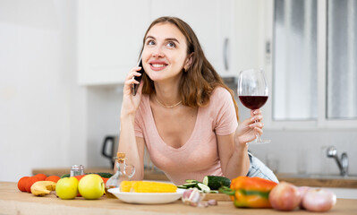 Wall Mural - Positive young woman with a glass of wine in her hands in the home kitchen communicates with someone on a mobile phone