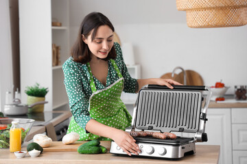 Canvas Print - Beautiful young woman cooking delicious sausages on modern electric grill in kitchen