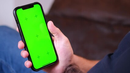 Unrecognizable young man sitting on the couch in a living room and using his smartphone. Scrolling, swiping, touching on a green screen with tracking markers. Browsing on the internet. App template. 
