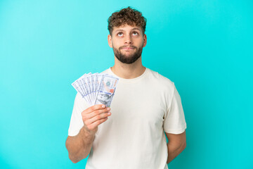 Canvas Print - Young caucasian man taking a lot of money isolated on blue background and looking up