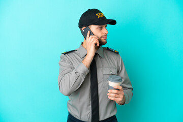 Wall Mural - Young security man isolated on blue background holding coffee to take away and a mobile