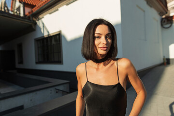 Wall Mural - Street summer sunny female portrait of a beautiful stylish fresh woman in a fashion black dress with spaghetti straps walking in the city in sunlight. Pretty chic girl at sunset