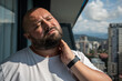 Bearded man in hot summer sun on balcony. Bald, unsmiling guy closed eyes in displeasure discontent from discomfort, holding on to sore neck, kneading ligaments, suffering from protrusion, pain, ache