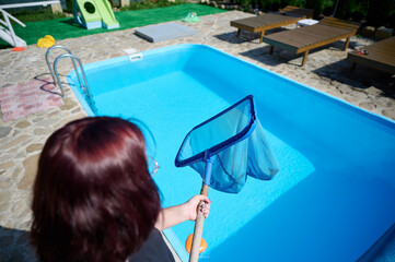 Wall Mural - Young woman cleans swimming pool. Personnel cleaning the pool from leaves in sunny summer day. Hotel staff worker cleaning the pool. Cleaning swimming pool service. Purification with a net. Close up