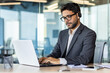 © Liubomir - A young Indian businessman is working in the office on a laptop and wearing headphones. Concentrated typing on the keyboard, chats.