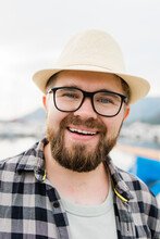 Close-up Handsome Millennial Man Wearing Hat And Glasses Near Marina With Yachts. Portrait Of Laughing Man With Sea Port Background