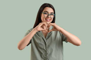 Wall Mural - Beautiful young woman wearing glasses and making heart with her hands on color background