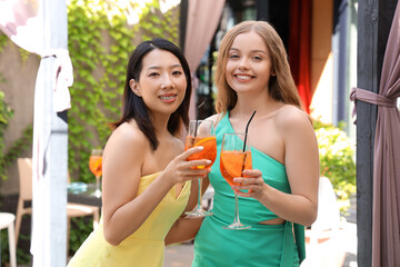 Wall Mural - Beautiful young women with glasses of tasty aperol spritz cocktail spending time in cafe, outdoors