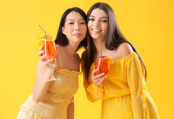 Wall Mural - Beautiful young women with glasses of tasty aperol spritz cocktail on yellow background