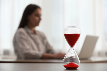 Wall Mural - Hourglass with red flowing sand on table. Woman using laptop indoors, selective focus