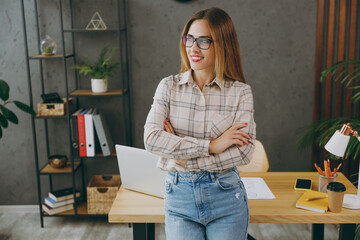 Wall Mural - Young successful smiling fun employee business woman wear shirt casual clothes glasses work at office sit on desk with pc laptop, hold hands crossed folded look aside. Achievement career job concept.