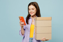 Young Happy Woman She Wears Purple Shirt Yellow T-shirt Casual Clothes Hold Stack Cardboard Blank Boxes Use Mobile Cell Phone Isolated On Plain Pastel Light Blue Background Studio. Lifestyle Concept.