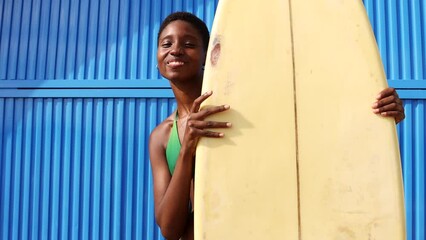 Wall Mural - Portrait of a surfer standing, in front of a blue background in swimwear, with her surfboard by her side. Happy woman doing beach activities during summer vacation.