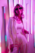 Leinwandbild Motiv Young beautiful hipster woman in street style fashion concept. Hot model wearing white suit and sunglasses. Female posing in pink neon light in studio interior