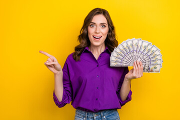 Sticker - Photo of impressed excited woman with curly hairdo dressed purple shirt hold dollars indicating empty space isolated on yellow background