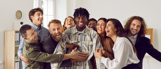 Happy diverse business team celebrating colleague's success together. Group portrait of cheerful joyful excited exhilarated friends hugging and congratulating young African American man on promotion