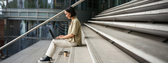 Wall Mural - Profile portrait of young asian woman with laptop, girl student sits on stairs outside building and types on computer, drinks takeaway coffee