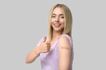 Canvas Print - Young woman with medical patch on arm showing thumb-up against grey background. Vaccination concept