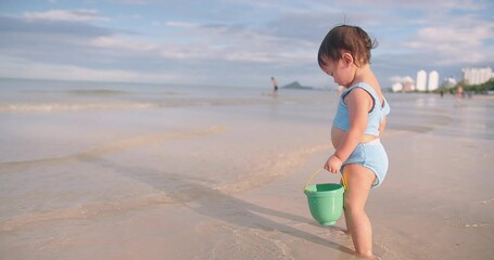 Wall Mural - Happy Cute Little Asian Toddler Child Girl in swimsuit Enjoying Vacation holding toy buckets playing sand on the beach with Waves Breaking on Seashore in summertime