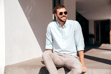 Handsome smiling stylish hipster lambersexual model. Modern man dressed in white shirt and trousers. Fashion male posing in the street urban background in sunglasses. Outdoors, sitting