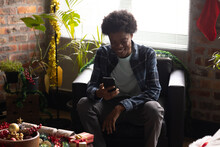 Happy African American Man Using Smartphone Sitting In Armchair At Home At Christmas Time