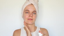 Confident young woman in white towel on head applies moisturizer cream on her face doing daily beauty care cosmetic routine after morning shower isolated over white background.