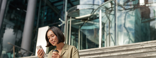 Wall Mural - Beautiful smiling asian girl, drinking coffee, using mobile phone and sits on stairs outside. Young woman video chat outdoors