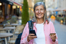 Portrait Of A Smiling Senior Woman Standing On A City Street, Holding A Credit Card And A Mobile Phone, Looking At The Camera.