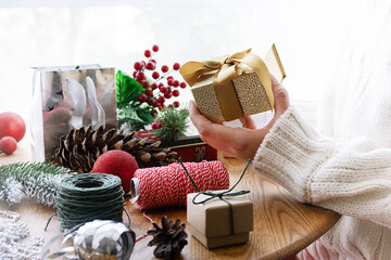 Wall Mural - Woman in warm woolen sweater holding golden Christmas gift box in her hand on the background of dofferent bright festive decorations close up.