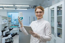 Portrait Of A Smiling Young Female Scientist Working In A Laboratory, Standing With A Tablet And A Flask With A Research Liquid In Her Hands. Smiling At The Camera.