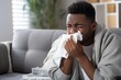 © NEM - Unhealthy young African American man in blanket suffering from flu blowing nose with napkin on sofa