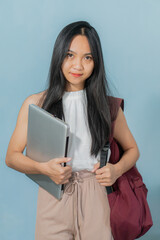Wall Mural - Portrait of a pretty cheerful casual Asian girl carrying backpack standing isolated over light blue background, holding laptop computer
