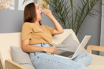 Wall Mural - Information browsing. Workspace research. Sleepy tired brown haired woman wearing beige T-shirt and jeans sitting on sofa and working on laptop and yawning needs rest posing in living room