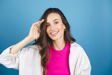 Wall Mural - Lovely coquettish brunette woman, winking playfully, flirting, throws her hair. Woman fix her hair. Brunette hair fashion model face smiling. Girl in pink top, white shirt isolated on blue background.