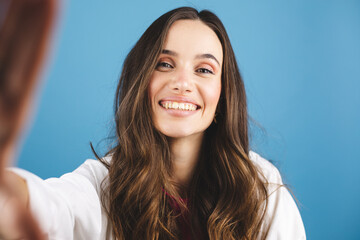 Wall Mural - Portrait of young happy brunette curly hair woman with toothy smile laughing isolated over blue wall background make selfie looking camera.