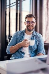 Wall Mural - Handsome smiling young man drinking coffee sitting at office desk and looking at camera