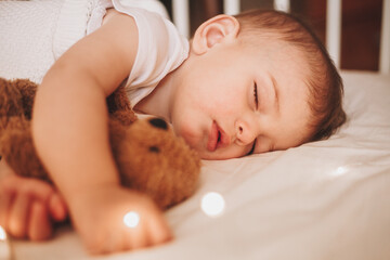 Wall Mural - A little baby in a white bodysuit sleeping in a white crib holding a teddy bear and surrounded by small lights