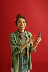 Wall Mural - Smiling woman is pointing on mobile phone while standing on red studio background