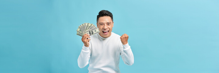 Wall Mural - Excited young handsome Asian man holding bunch of money banknotes isolated on blue banner